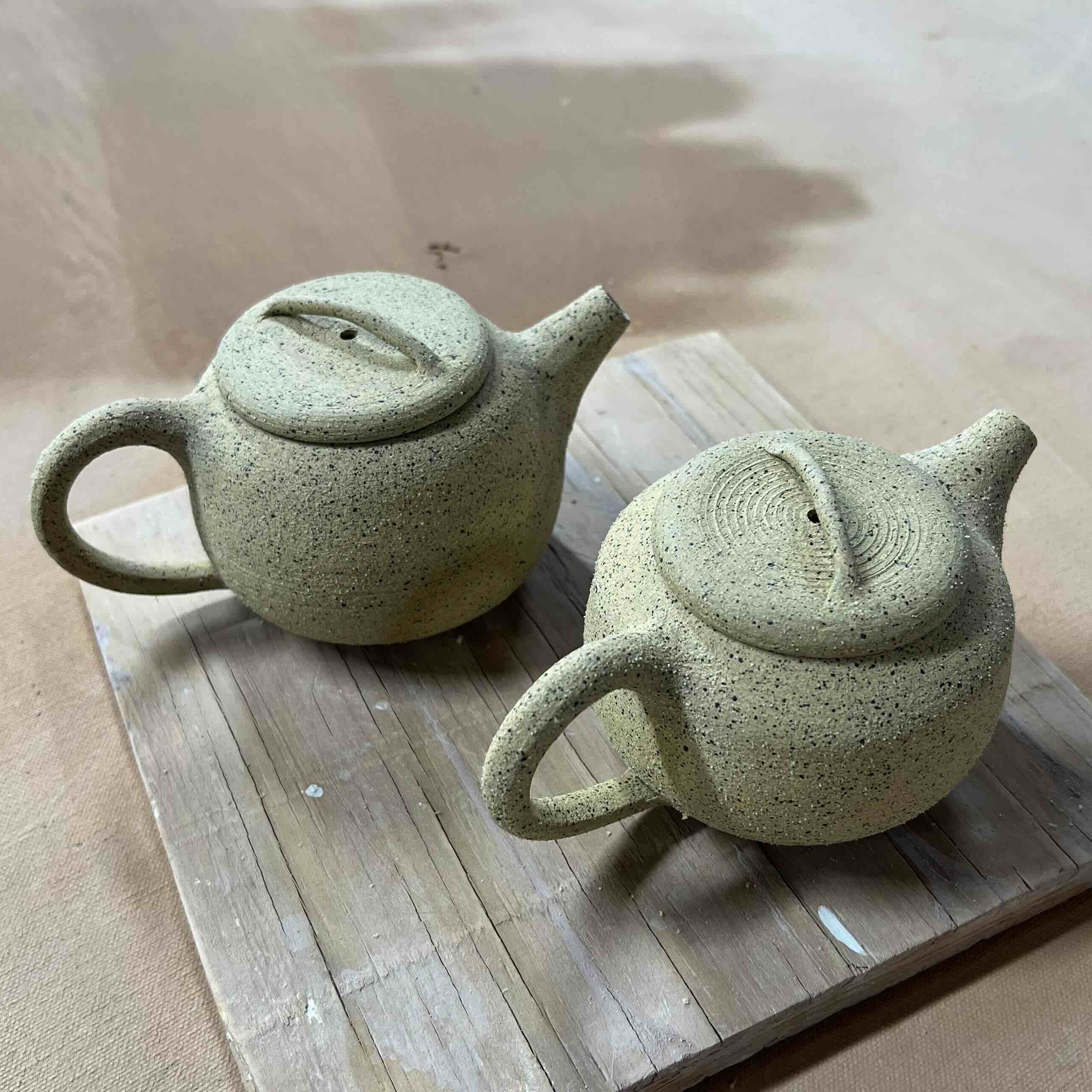 Two fired but unglazed teapots on a wooden square.